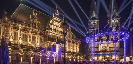 Der beleuchtete Bremer Dom und das beleuchtete Bremer Rathaus bei Nacht. Davor strahlen zahlreiche Scheinwerfer in den Himmel.