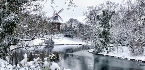 Eine eingeschneite Winterlandschaft ("Wallanlagen") mit einem zugefrorenem Wasserlauf. Im Hintergrund steht die eingeschneite "Mühle am Wall".