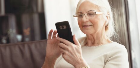 Eine ältere Frau sitzt auf einem Sofa und nutzt ein Smartphone.