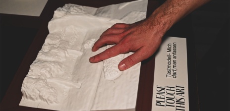 Eine Hand ertastet ein Tastmodell. Darunter ist ein Schild mit der Aufschrift: "Please touch this art. Tastmodell - mich darf man anfassen."