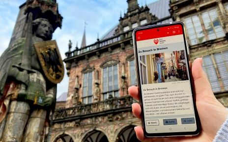 Jemand hält ein Smartphone vor dem Rathaus in Bremen. Auf dem Bildschirm sieht man die Startseite von "Dein Bremen Guide".
