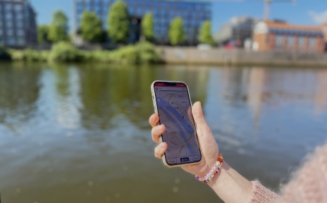 Das Bild zeigt ein Hand, welche ein Handy hält auf der Dein Bremen Guide geöffnet ist. Im Hintergrund sieht man die Weser.