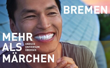 Ein lachender Mann vor dem Science Center Universum Bremen. Er hat schwarze Haare. Schriftzug im Bild: "Bremen - Mehr als Märchen. Unesco, Universum, Übersee". 