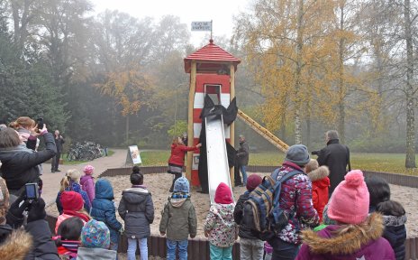 Auf dem Bild ist ein Spielplatz in einem Park zu sehen. Senatorin Anja Stahmann enthüllt ein Spielgerät in Form eines Leuchtturms. Im Publikum sind viele Kinder zu sehen. Das Spielgerät ist mit Klettermöglichkeiten und Rutsche ausgestattet. Auf dem Dach befindet sich eine Fahne mit der Aufschrift "Platz der Kinderrechte".