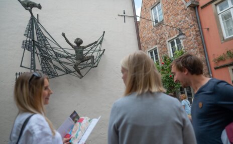 Zwei Frauen und ein Mann stehen vor der Ottjen-Alldag-Plastik im Schnoor und unterhalten sich. 