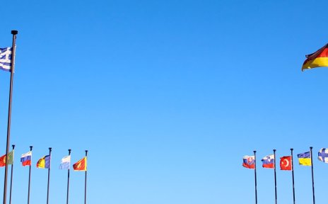 Unterschiedlichste Fahnen aus verschiedenen Ländern wehen im Wind vor blauem Himmel. Sie sind an Maste im Kreis arrangiert.