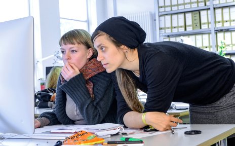 Zwei junge Frauen am Schreibtisch schauen auf einen Monitor.