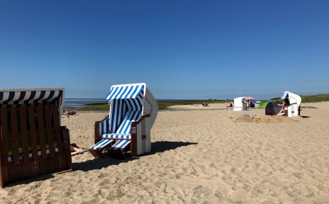 Weißer Sandstrand mit Strandkörben und blauem Himmel. Im Hintergrund sind Wiesen und das Meer zu sehen.