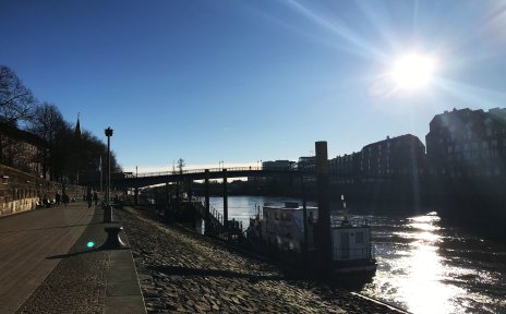 Zu sehen ist ein gepflasterter Fußweg, auf dem Personen spazieren gehen. Schräg abfallende Steine führen zum Wasser. Auf dem Wasser, das rechts im Bild zu sehen ist, liegt ein Schiff. Im hinteren Teil des Bildes ist eine Brücke zu erkennen, die über das Wasser führt.
