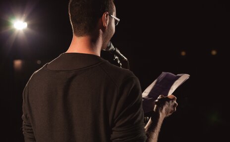 Ein Mann von hinten, vor ihm ein Mikrophon. Er liest aus einem Buch vor.