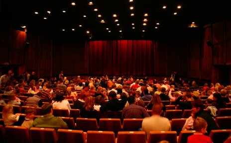 Man sieht einen in rot gestalteten Theatersaal, in dem Menschensitzen. Man blickt auf die Bühne, die von einem roten Vorhang verhangen ist. Der Raum ist in warmes Licht getaucht.