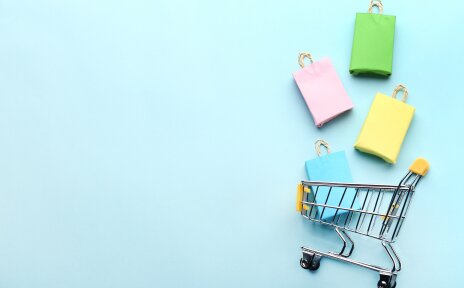 Über einem kleinen Einkaufswagen schweben gebastelte Einkaufstüten. Der Hintergrund ist türkis.