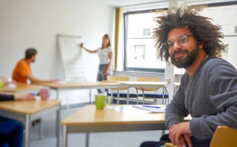 Ein Mann mit wuschelig-lockigem Haar und einer Brille sitz in einem Kursraum und lächelt in die Kamera. Im Hintergrund andere Kursteilnehmende und eine Lehrerin.