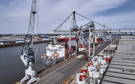 Zu sehen ist der Neustädter Hafen. Ein großes Containerschiff hat angelegt. Zudem sind einige Kräne, das Wasser und eine Lagerhalle zu sehen. In einiger Entfernung befinden sich noch zwei weitere Containerschiffe. 