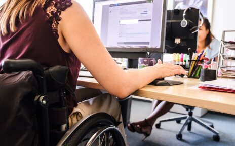 Eine Frau im Rollstuhl sitzt an einem Schreibtisch in einem Büro. Sie arbeitet am Computer.