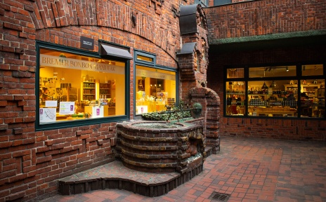 Außenansicht der Bremer Bonbonmanufaktur im Handwerkerhof mit beleuchteten Schaufenstern und Backsteinarchitektur.