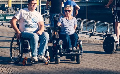 Ein lächelnder Mann und eine Frau im Rollstuhl an der Schlachte. Außerdem sind Menschen auf Segways unterwegs.