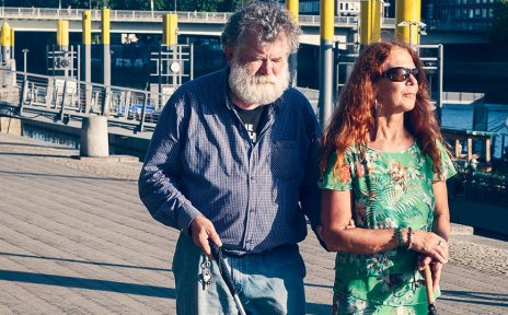 Ein Mann und eine Frau, beide mit Blindenstock, gehen an einem Fluss spazieren. Im Hintergrund sind Schiffe und eine Brücke zu sehen.