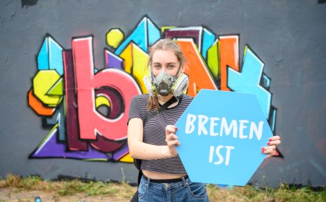 Eine Frau mit Atemmaske steht vor einem bunten Graffiti. In der Hand hält sie ein Schild mit "Bremen ist".