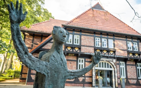 Bronzeskulptur mit ausgebreiteten Armen vor einem historischen Fachwerkgebäude im Grünen.