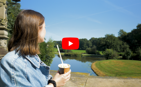 Eine Frau steht mit dem Rücken zum Betrachtenden auf der Melchersbrücke im Bürgerpark und blickt auf grüne Wiesen und Wasser. In der Hand hält sie einen Kaffeebecher mit Strohhalm. In der Mitte befindet sich das YouTube-Logo: ein weißes Dreieck auf einem roten Rechteck.