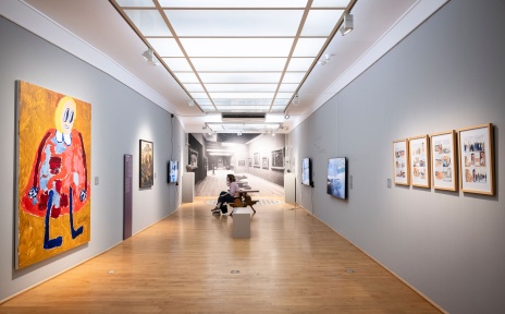 Eine Person sitzt auf einer Holzbank in einem Ausstellungsraum und schaut auf einen Bildschirm an der Wand. 
