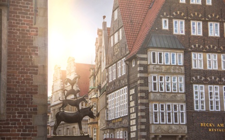 Im linken Teil des Bildes sind die Bremer Stadtmusikanten zu sehen. Im rechten Teil ein historisches Gebäude.