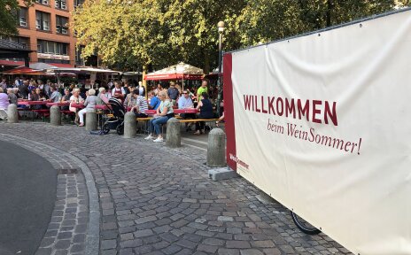 Viele Menschen sitzen draußen an langen Tischen auf einem Platz. Daneben ist ein großes Schild aufgehängt mit der Aufschrift: Willkommen beim WeinSommer!