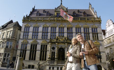 Eine Frau und ein Mann stehen vor einem Gebäude, an dem eine rote, weiße Fahne weht. 