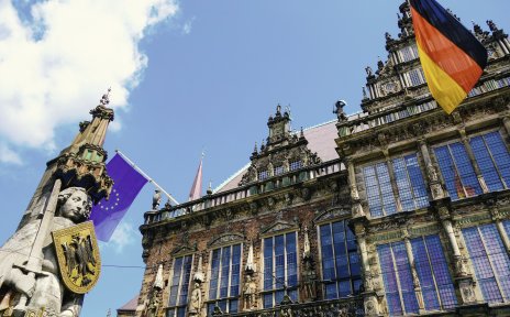 Froschperspektive auf eine steinerne Statue, die ein Schild und ein Schwert hält und auf ein Gebäude, an dem die Flagge Deutschlands und Europas hängt. 