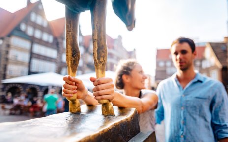Eine Frau fasst dem Esel der Bremer Stadtmusikanten um die Füße. Sie lächelt dabei einen Mann an. 