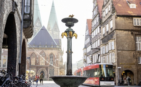 Ein goldverzierter Brunnen in einer Bremer Einkaufsstraße. Im Hintergrund ist der Bremer Dom und eine Straßenbahn zu sehen.