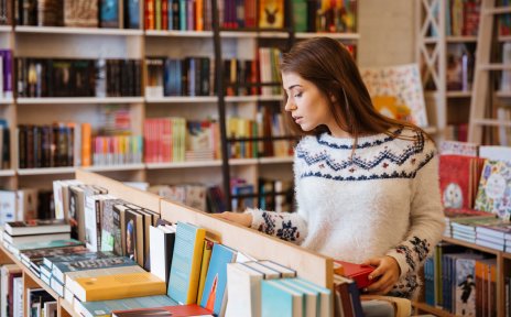 A young woman is standing in front of a table-high shelf in a bookshop, looking at the books. In the background are large bookshelves.