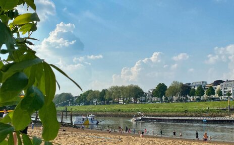 Ausblick auf den Strand am Café Sand, auf der Weser die Sielwallfähre und ein Binnenschiff, am Strand Menschen in Badekleidung, am linken Bildrand grüne Blätter eines Baumes