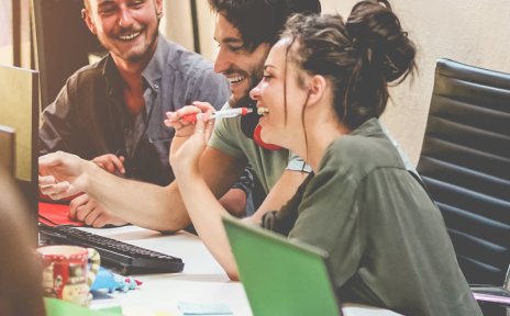 Junge, gut gelaunte Leute sitzen an einem großen Besprechungstisch und tauschen sich aus. Auf dem Tisch stehen Laptops und andere Arbeitsutensilien.