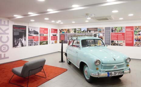Ein hellblauer Oldtimer steht in einem weißen Raum. An den Wänden hängen Tafeln mit Bildern und Texten und der Schriftzug Focke ist zu lesen.
