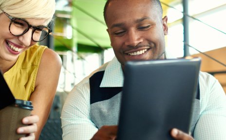 Zwei lachende Personen, eine Frau und ein Mann, sitzen auf einer Treppe. Sie hat ein Notizbuch in der Hand, er zeigt etwas auf einem Tablet.
