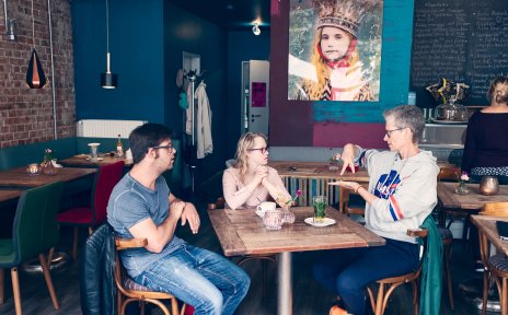 Zwei Frauen und ein Mann sitzen an einem Tisch in einem Café. Eine der Frauen spricht in Gebärdensprache.