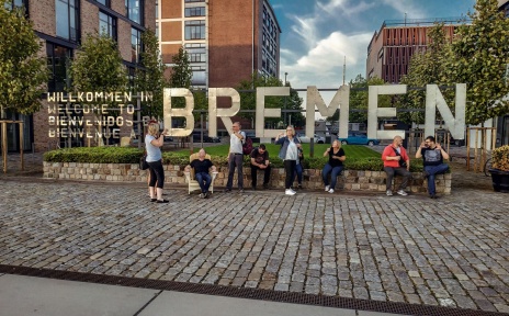 Menschen stehen und sitzen vor einer Skulptur aus Metall mit der Aufschrift "Willkommen in Bremen". Sie unterhalten sich in Gebärdensprache. 