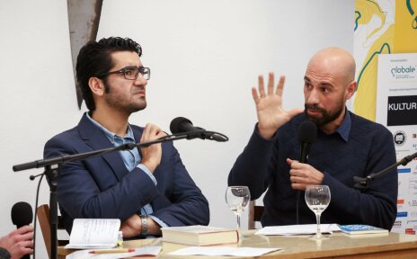 Zwei Männer reden miteinander vor ihnen steht ein Mikrofon, Der Linke Mann hat dunkle Haare und trägt eine Brille. Der Rechte Mann hat eine Glatze und einen Bart.