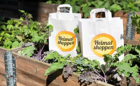 Man sieht zwei Papiertüten mit dem Logo der Aktion "Heimat Shoppen", die in einem Beet stehen.