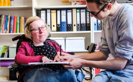 Eine Frau sitzt in einem Rollstuhl mit Rollstuhltisch. Neben ihr sitzt ein Mann und schreibt etwas auf ein Papier, das auf dem Rollstuhltisch liegt. Im Hintergrund sind Regale mit Aktenordnern und Büchern zu sehen.