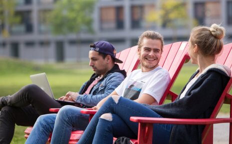 Drei Studierende, eine blone Frau mit Dutt, ein blonder Mann und ein brünetter Mann mit Baseballkappe, sitzen auf roten Holzstühlen auf dem Campus der Universität Bremen. Sie reden miteinander und lachen. Der Mann mit der Kappe schaut auf seinen Laptop.