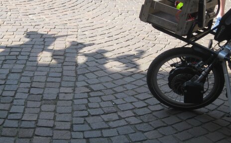 Vorderreifen eines Handbikes. Der Boden ist mit Kopfsteinpflaster versehen, man sieht einen Schatten der Bremer Stadtmusikanten.