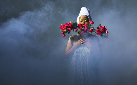 In Nebel gehüllt steht eine weiß gekleidete Person. Sie trägt eine goldenen Maske und hat einen Strauß pinker Blumen in der Hand.