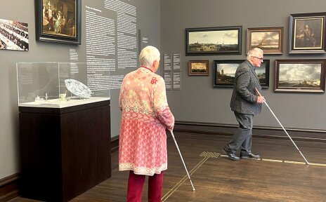 Zwei Personen laufen in einem Ausstellungsraum der Kunsthalle Bremen. Sie orientieren sich mit Taststöcken am Bodenleitsystem. An der Wand sind Gemälde, in einer Glasvitrine ist Porzellan ausgestellt.