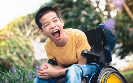 Ein lachender Junge im Rollstuhl. Er befindet sich draußen in der Natur und hält eine lila Blüte in der Hand.