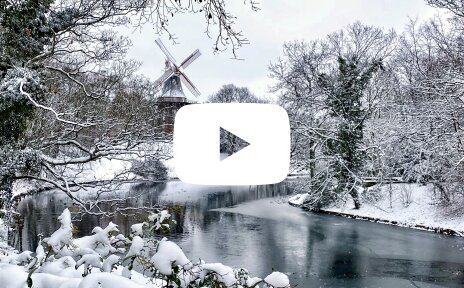 In der Mitte des Bildes befindet sich ein weißes Icon mit einem transparenten Dreieck in der Mitte. Im Hintergrund ist eine verschneite Schneelandschaft zu erkennen mit der "Mühle am wall".