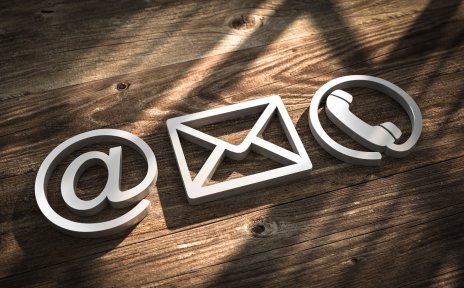 Ein @-Zeichen, ein Briefsymbol und ein Telefonhöhrer-Symbol vor einem hölzernen Hintergrund