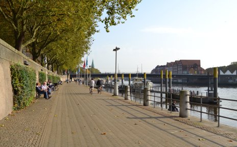 Eine Promenade mit Menschen am Fluss, im Hintergrund eine Brücke, die über den Fluss führt.